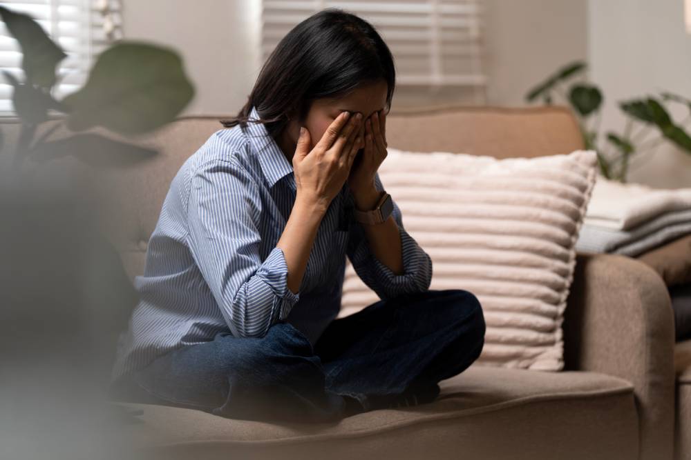 2764194505 A woman sits on a couch, her hands covering her face.